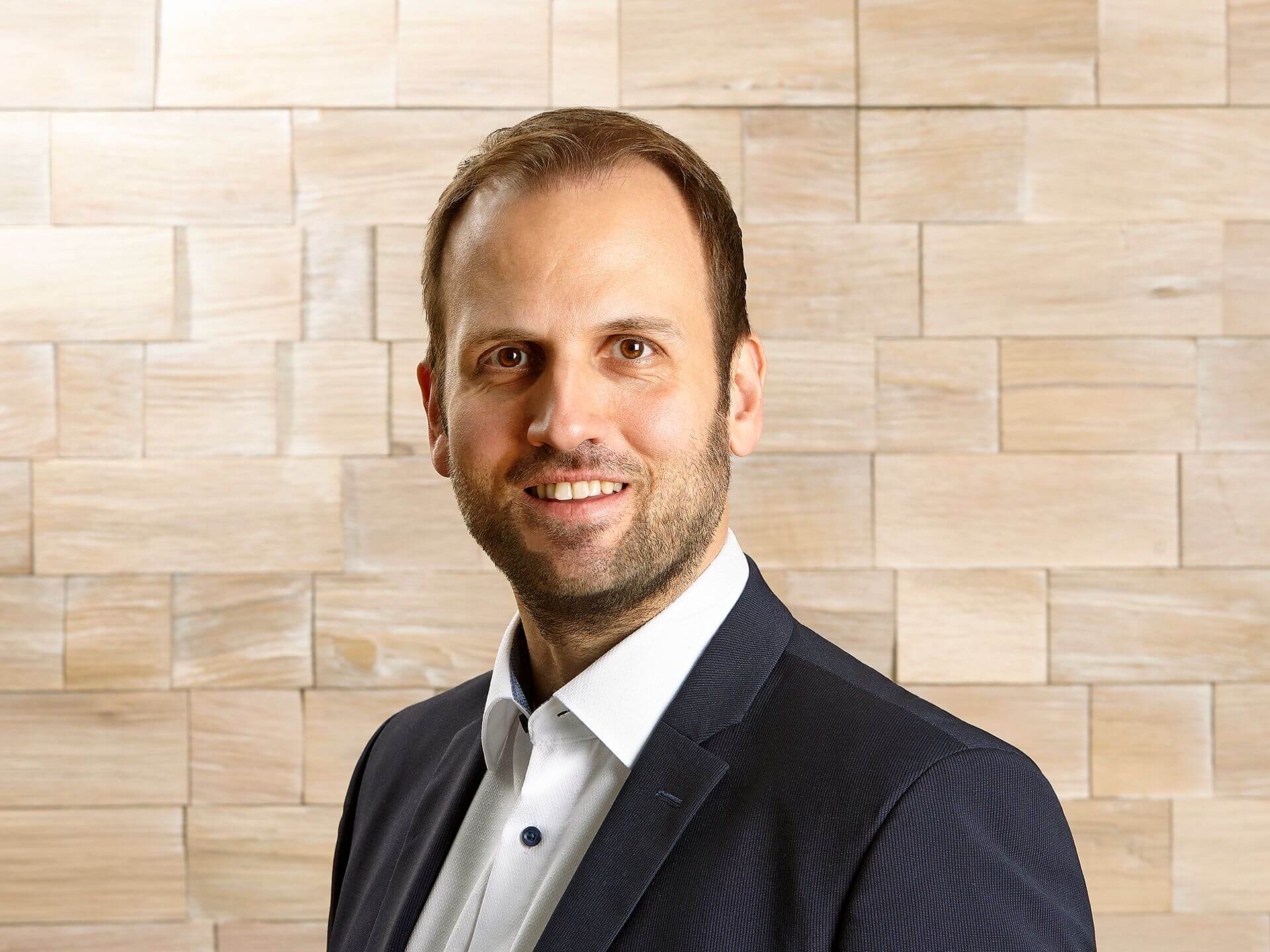 Christian Zoehrlaut, lächelnder Mann mit Bart und dunklem Anzug vor einer Wand aus Holzfliesen