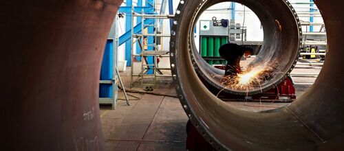 Person in Overall und Sicherheitsausrüstung, die in einer Fabrik ein großes Metallrohr schweißt