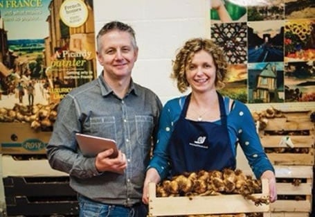 Man holding a tablet standing beside a woman holding a crate of ginger
