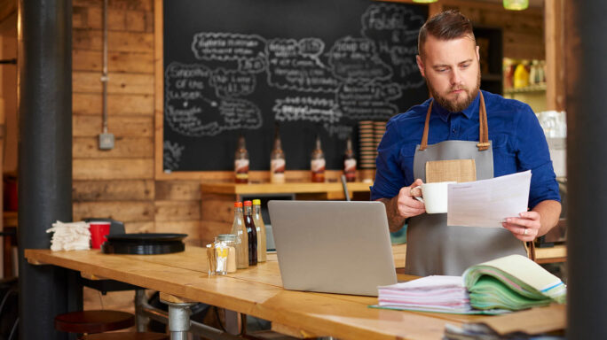 coffee shop owner look at invoices in front of laptop standing u drinking coffee