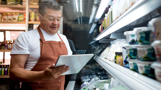 man in apron with an ipad stands in front of a fridge