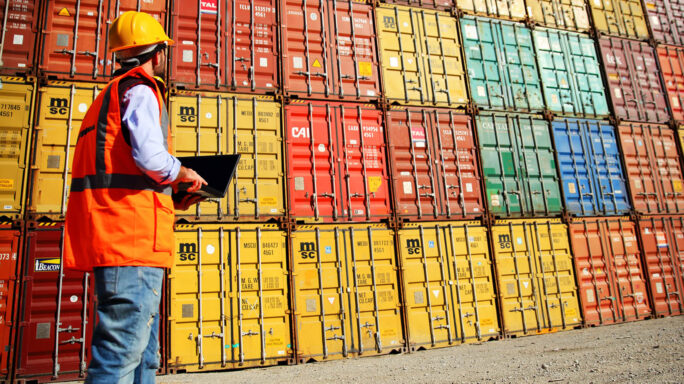 man in orange high vis vest looks at stacked shipping containers