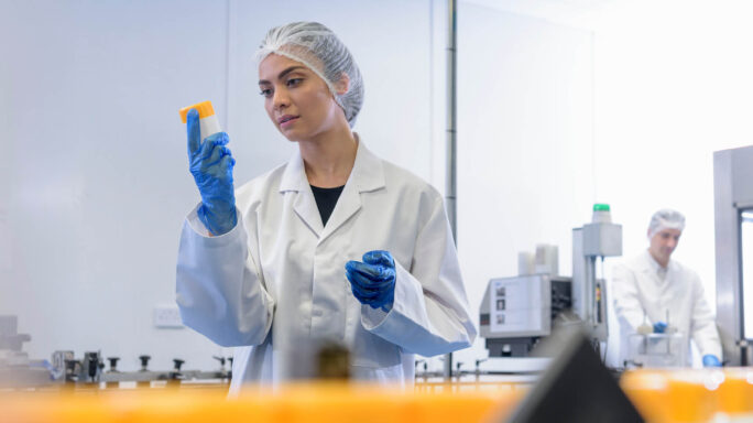 lab worker in white coat and blue gloves looks at sample in a container