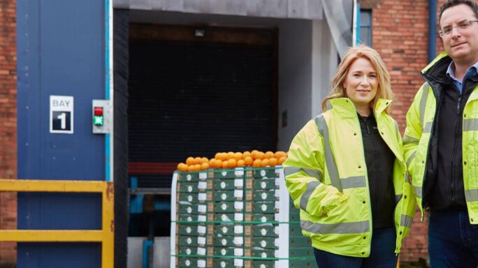 man and woman wearing high vis jackets in front of a warehouse with oranges