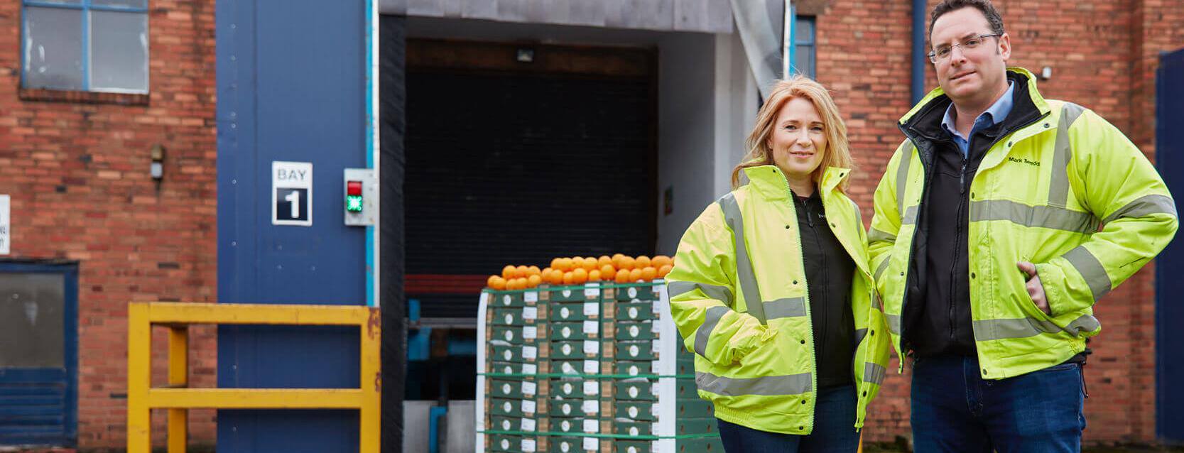 man and woman wearing high vis jackets in front of a warehouse with oranges