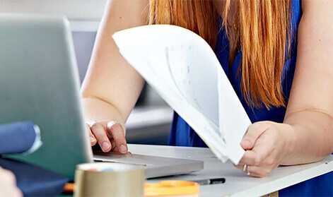 Woman's hands holding a report and typing on a laptop at a busy desk
