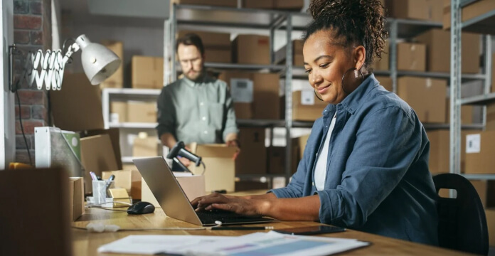  Person working on a laptop at a desk in a warehouse, with another person standing in the background among shelving units and boxes