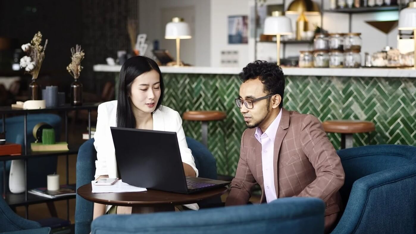  Two people reviewing information on a laptop while seated in a café or workspace