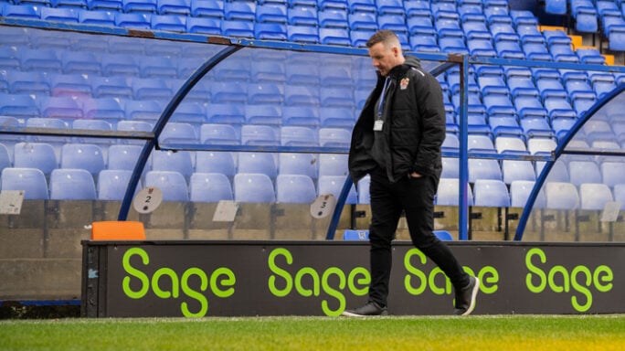 A man walks across the on-field dugout of a football pitch. Sage promotional banners flank the background.