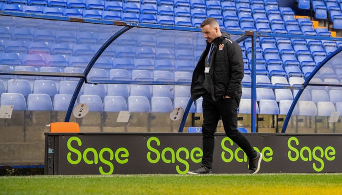 A man walks across the on-field dugout of a football pitch. Sage promotional banners flank the background.