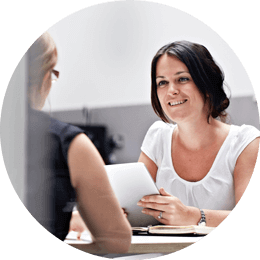 Roundel of a woman with dark hair wearing a white T-shirt talking to another woman at a laptop
