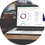 Roundel of a laptop displaying a budget planner, a notepad and a glass of soda on the screen positioned on a wooden counter in a bar