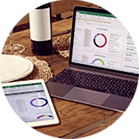 Roundel of a laptop and tablet displaying a budget planner on a wooden table set with plates, cutlery and a bottle of wine