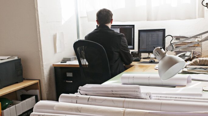 A man sits in front of a computer with the room full of files