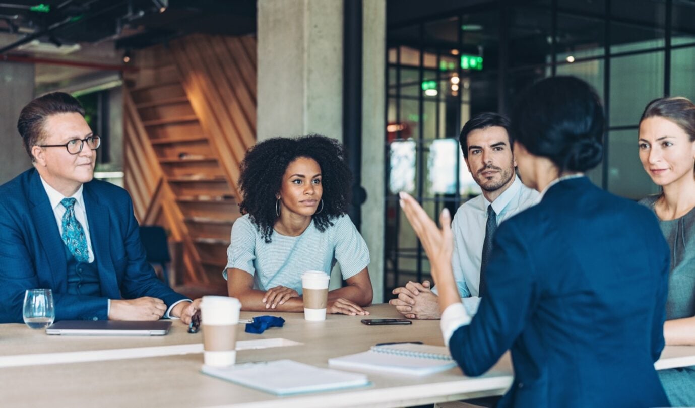 Reunión de trabajo en una sala de oficinas, con varias personas sentadas alrededor de una mesa mientras una persona realiza una presentación. Sobre la mesa hay ordenadores portátiles, documentos y materiales de trabajo, reflejando un entorno colaborativo y de toma de decisiones.
