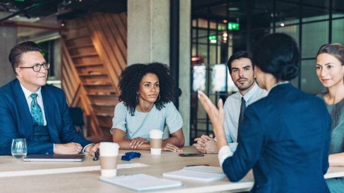 Reunión de trabajo en una sala de oficinas, con varias personas sentadas alrededor de una mesa mientras una persona realiza una presentación. Sobre la mesa hay ordenadores portátiles, documentos y materiales de trabajo, reflejando un entorno colaborativo y de toma de decisiones.