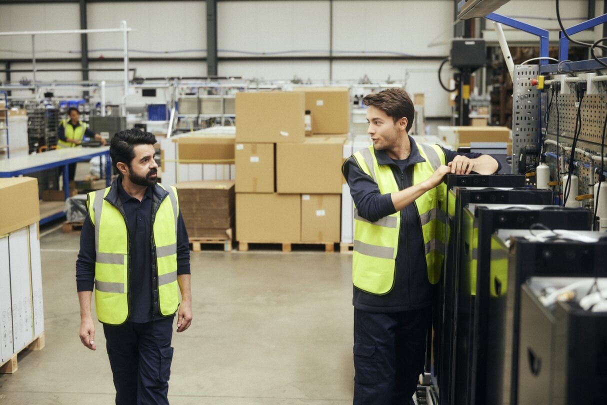 Personal de almacén con chalecos de alta visibilidad inspecciona mercancías y revisa documentación junto a estanterías con cajas y un montacargas en un entorno logístico.