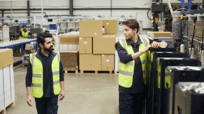 Personal de almacén con chalecos de alta visibilidad inspecciona mercancías y revisa documentación junto a estanterías con cajas y un montacargas en un entorno logístico.