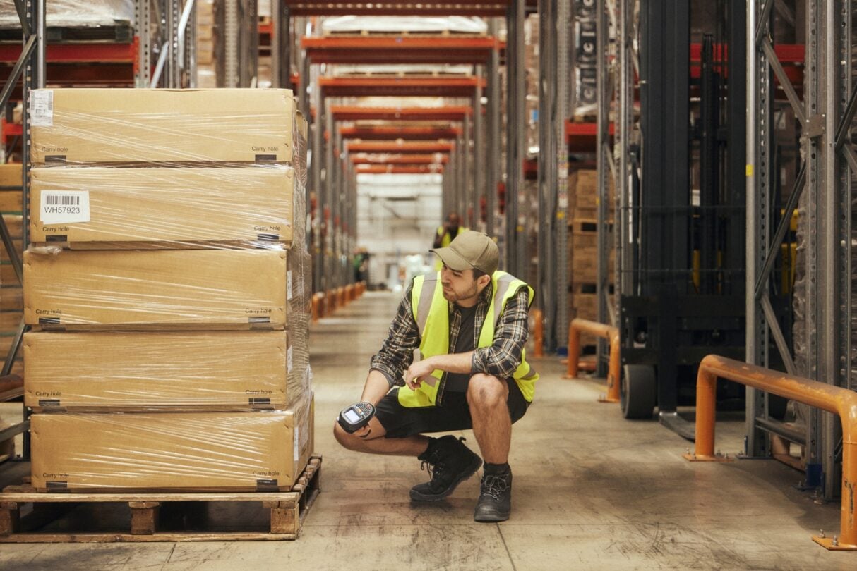 Trabajador en un almacén revisando mercancías junto a estanterías con palés y cajas, en un entorno logístico de distribución y almacenamiento.