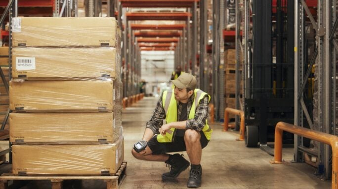 Trabajador en un almacén revisando mercancías junto a estanterías con palés y cajas, en un entorno logístico de distribución y almacenamiento.