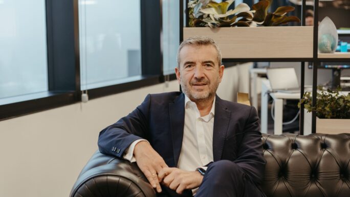 Un hombre de traje está sentado en un sofá de cuero negro y sonríe. Está en un espacio de oficina moderno con grandes ventanales y estanterías con plantas detrás de él.