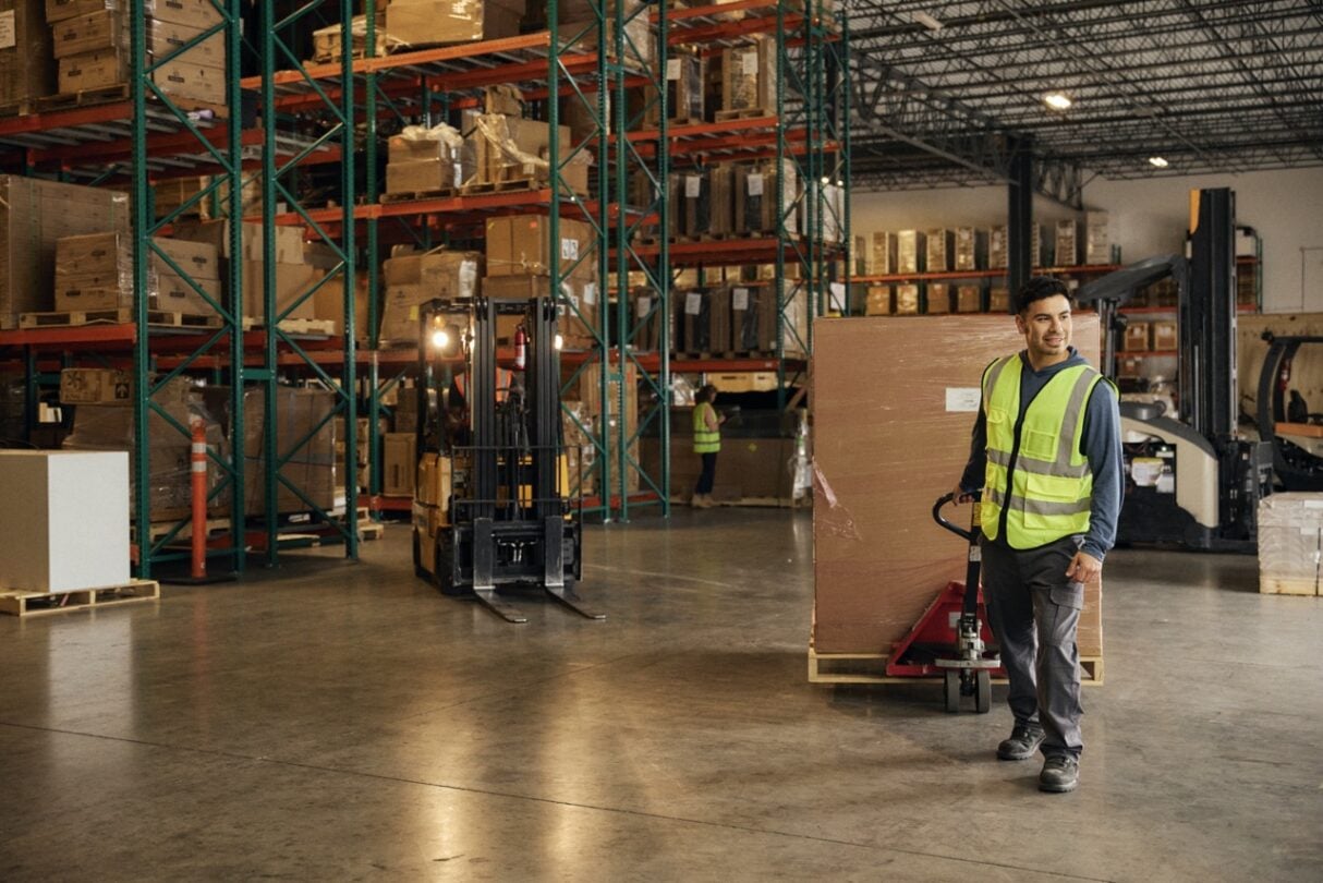Pessoa a mover uma caixa e um porta-paletes num armazém com estantes cheias de mercadorias.