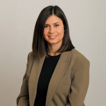 Retrato profesional de una mujer con el pelo liso y oscuro que le llega hasta los hombros, vestida con una chaqueta beige sobre un top negro, mirando al frente sobre un fondo liso de color gris claro.