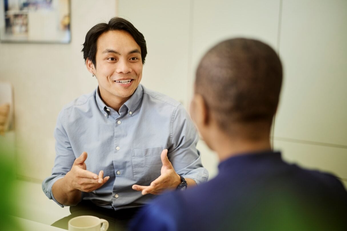 Persona conversando con un compañero durante una reunión en la oficina, gesticulando mientras comparte ideas en un entorno de trabajo colaborativo.