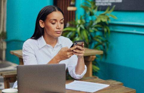  woman sitting outdoors looking at her phone with a laptop on the table in front of her