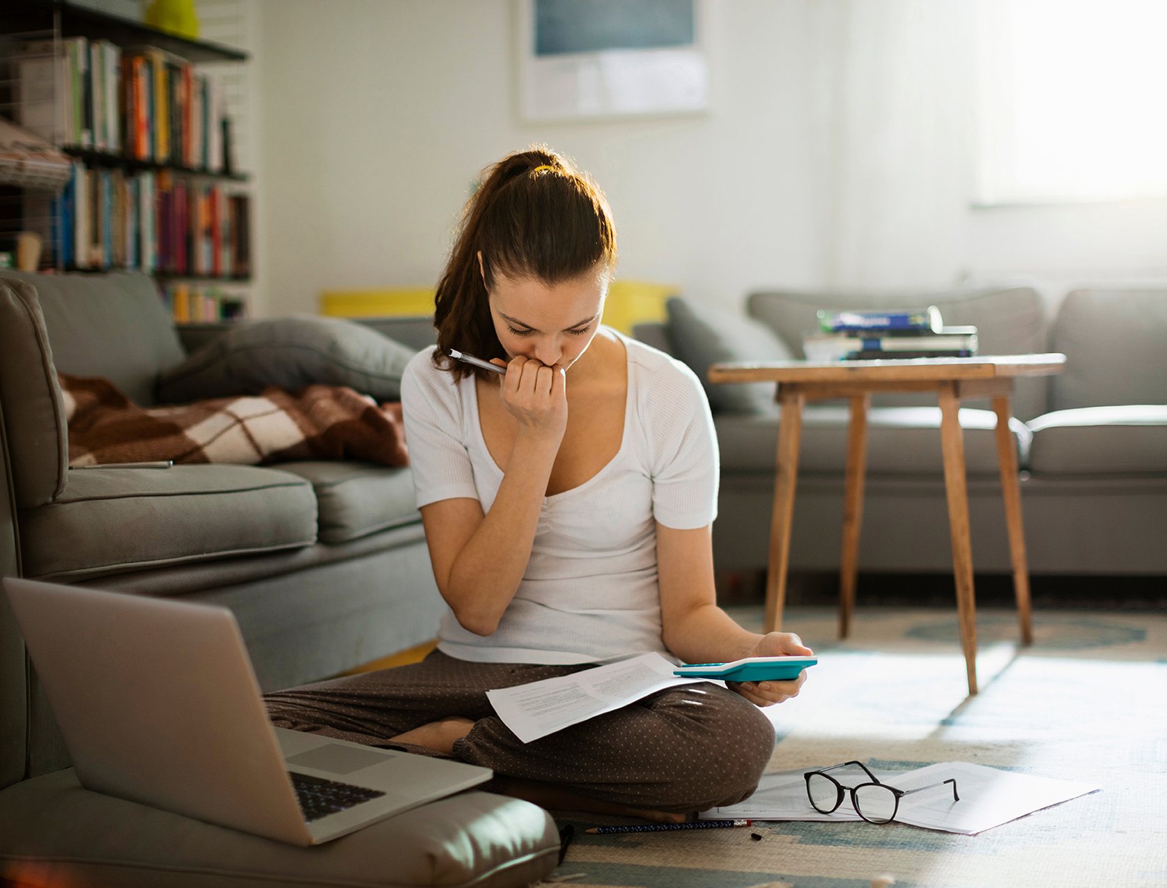 Een jonge vrouw zit op de vloer met haar laptop, verdiept in papierwerk