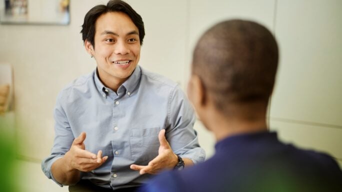 Pessoa a conversar com um colega durante uma reunião no escritório, gesticulando enquanto partilha ideias num ambiente de trabalho colaborativo.
