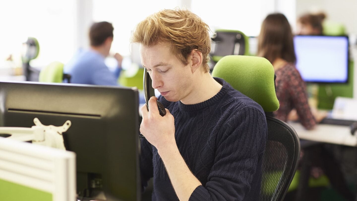 Pessoa sentada num escritório, a trabalhar num computador de secretária, com outros postos de trabalho e pessoas desfocadas ao fundo.