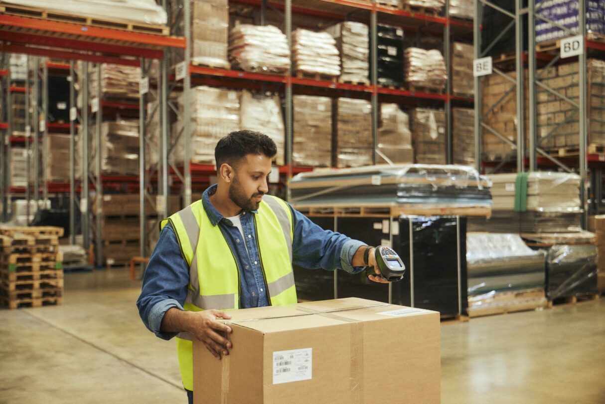 Trabalhador num armazém a manusear uma caixa de cartão, usando colete refletor e luvas. Ao fundo, prateleiras metálicas com paletes e materiais armazenados.