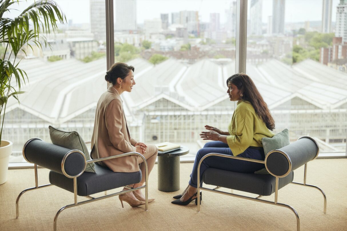 Uma mulher jovem, vestida com um blazer cinza e uma camisa branca, está sentada à frente de um computador portátil num ambiente de escritório moderno. Ela está a sorrir enquanto olha para o ecrã, sugerindo que está satisfeita com o que vê. Este cenário transmite uma atmosfera de produtividade e profissionalismo.