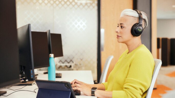 Trabalhador sentado à secretária durante uma reunião no escritório, com computador portátil aberto e documentos sobre a mesa.