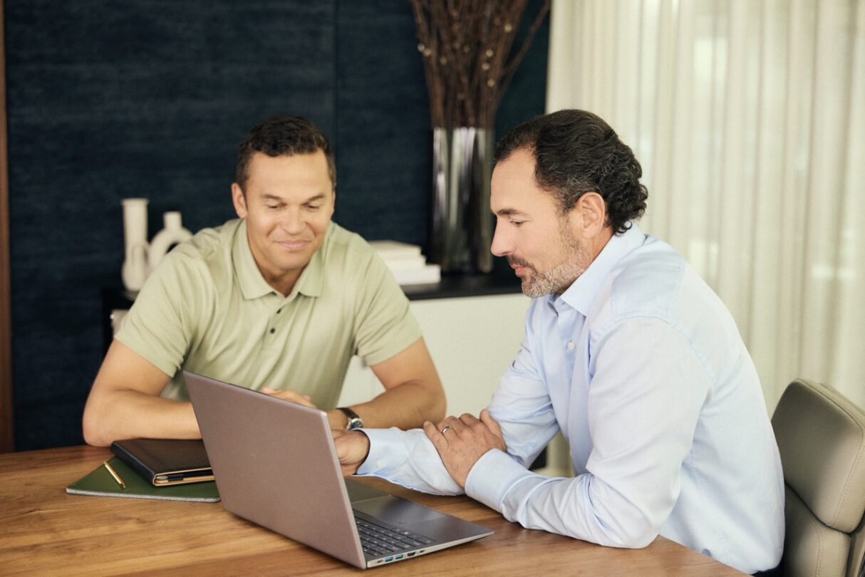 Funcionário sentado à mesa de reunião, concentrado, com documentos e portátil à frente, enquanto escuta atentamente os colegas à sua volta.
